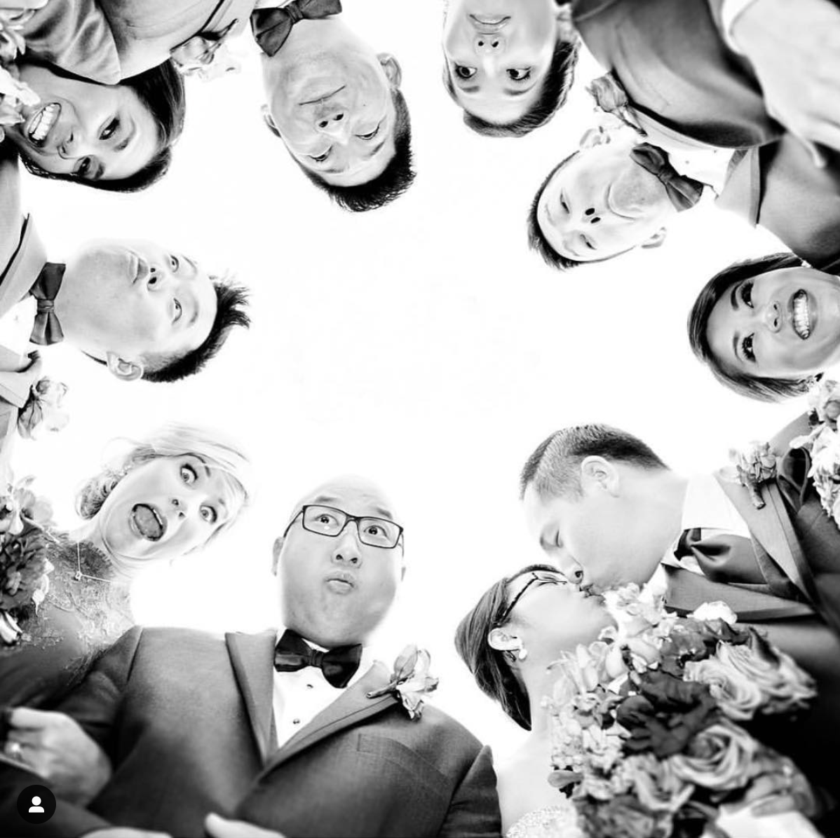 Low-angle shot of the groomsmen and bridesmaid posing in a circle and looking down at the camera alongside the bride and groom kissing