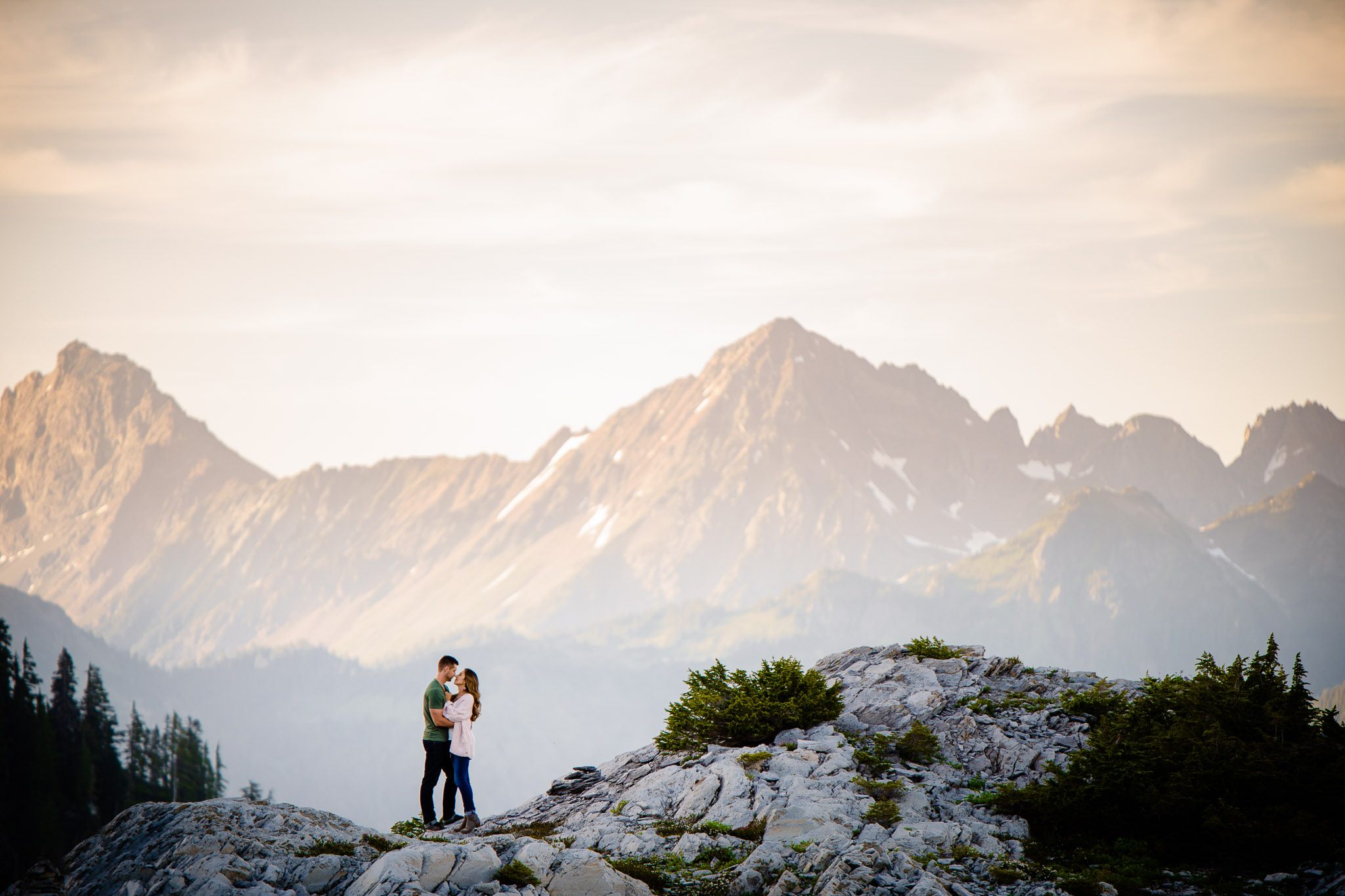 A couple holding each other for a pose on top of a cliff with mountain peaks in the background