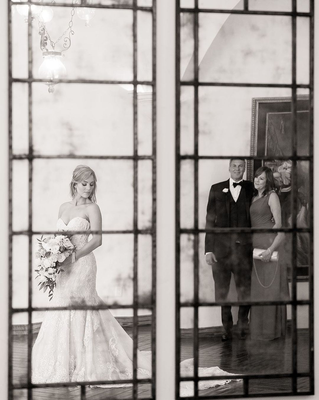 Black and white image from the POV of a window of a bride holding her bridal bouquet as her parents look emotionally from a distance  