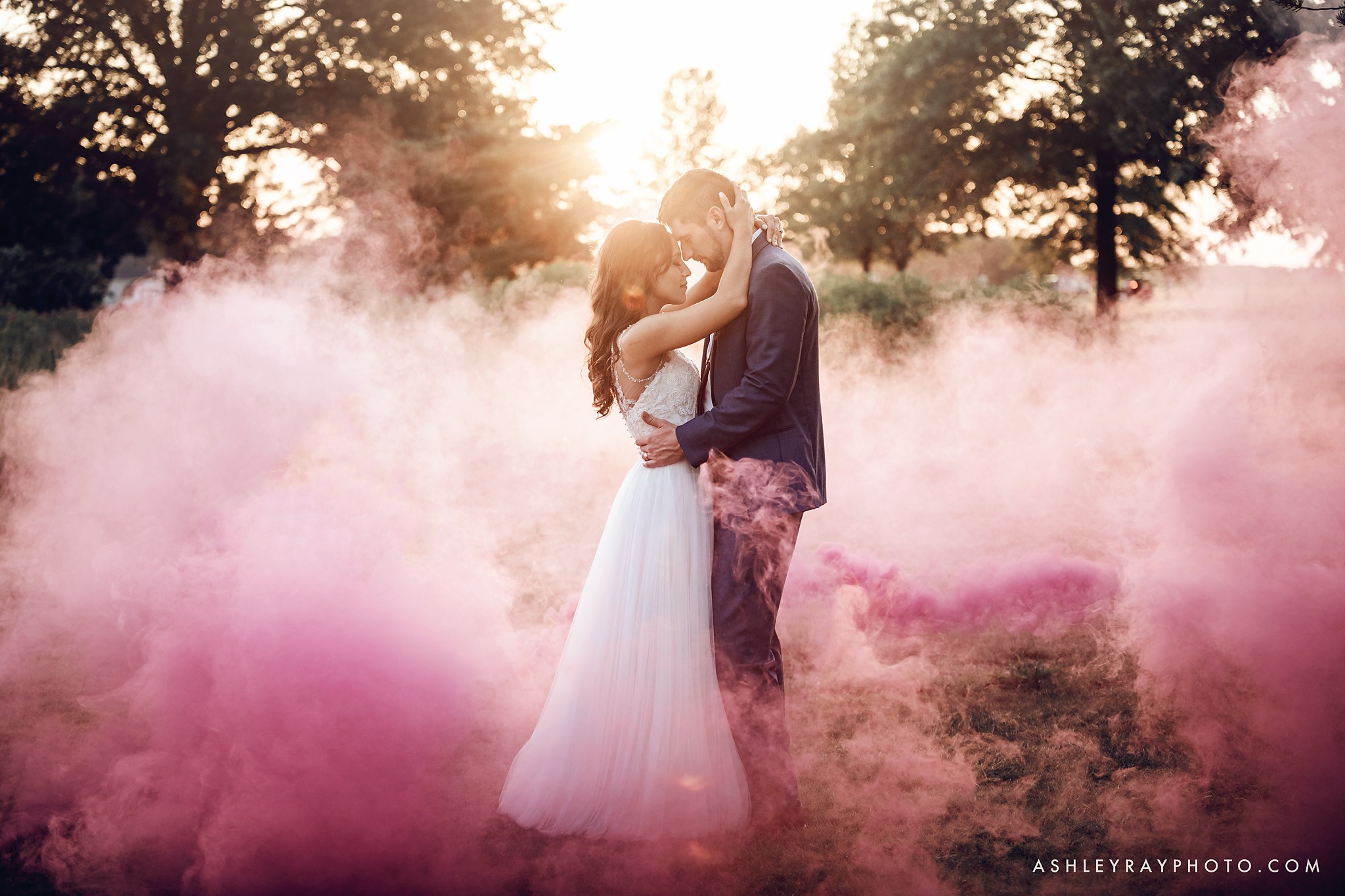 A bride and groom posing while holding each other as they are surrounded by pink color smoke bomb  