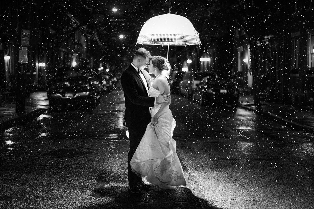 A black and white image of a bride and groom holding an umbrella as it rains