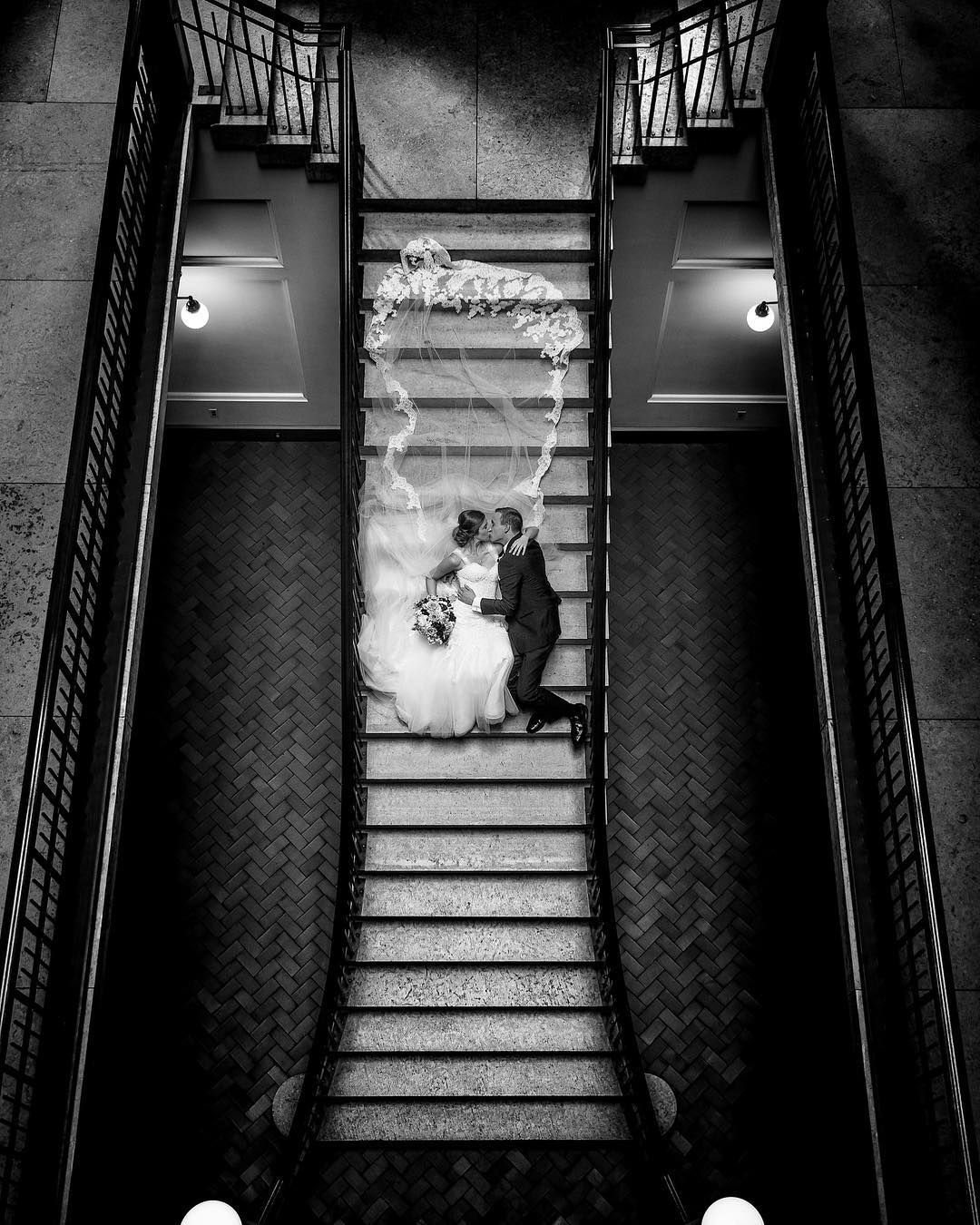 Bird’s-eye view of a bride and groom kissing and lying down on a staircase with the bridal veil spread out