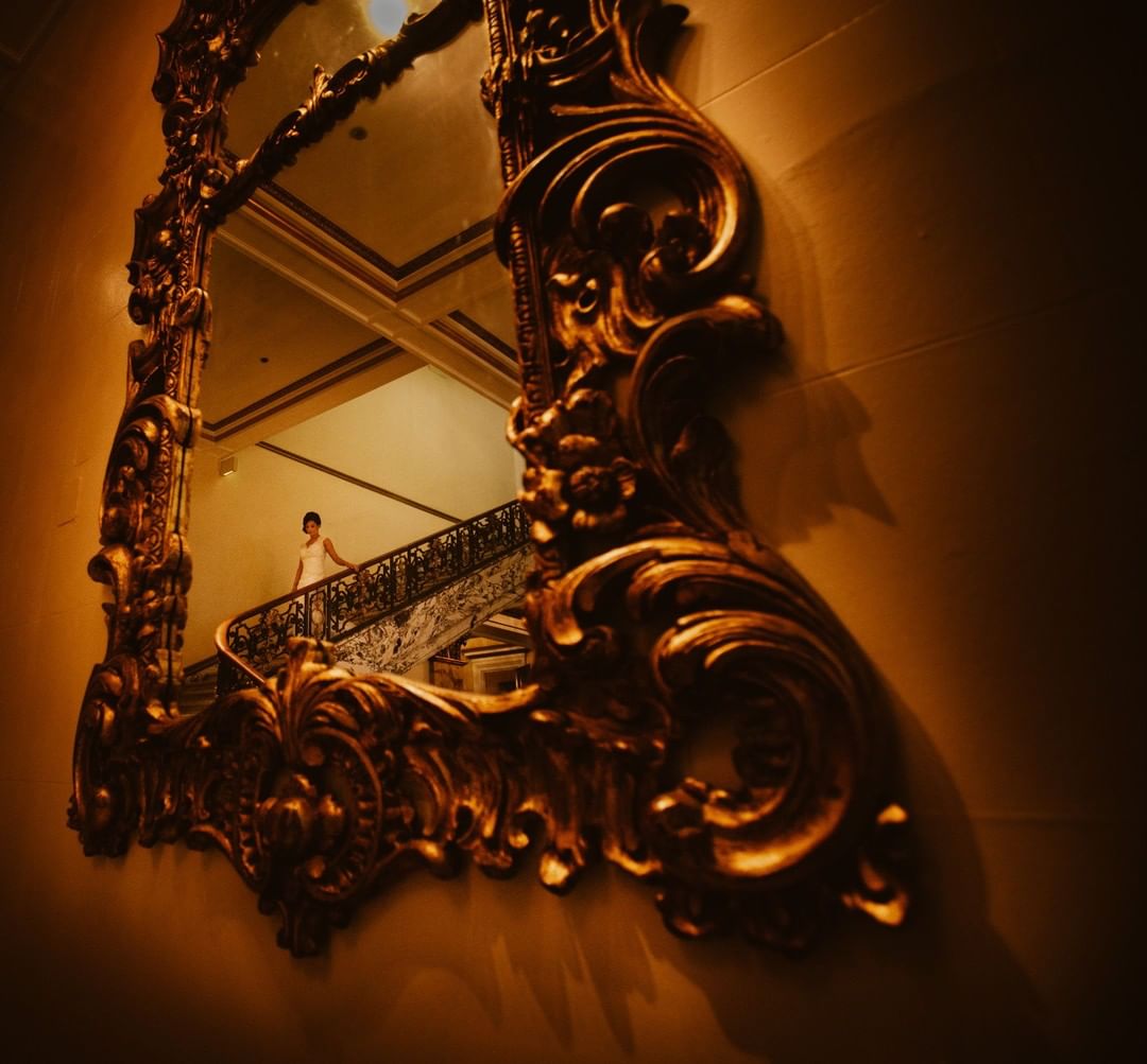 A reflection of a bride walking down the stairway in a golden framed mirror