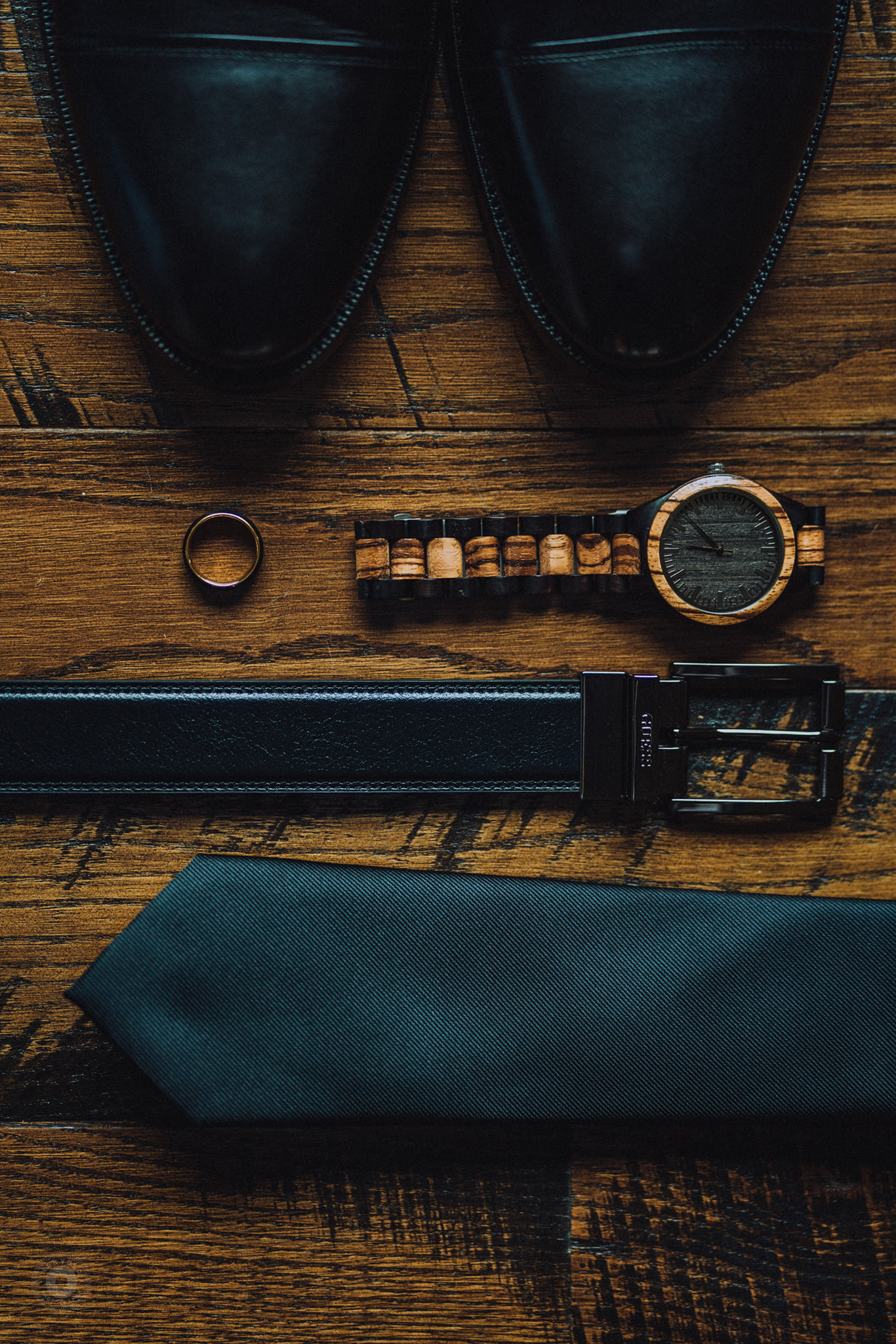 A close up shot of a watch, belt, shoes, a ring, and a tie placed on top of a wooden table