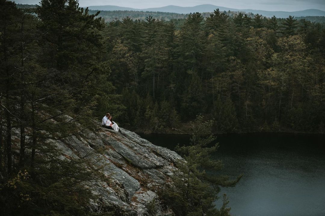 A couple sitting on top of a mountain cliff overlooking a lake amidst a forest
