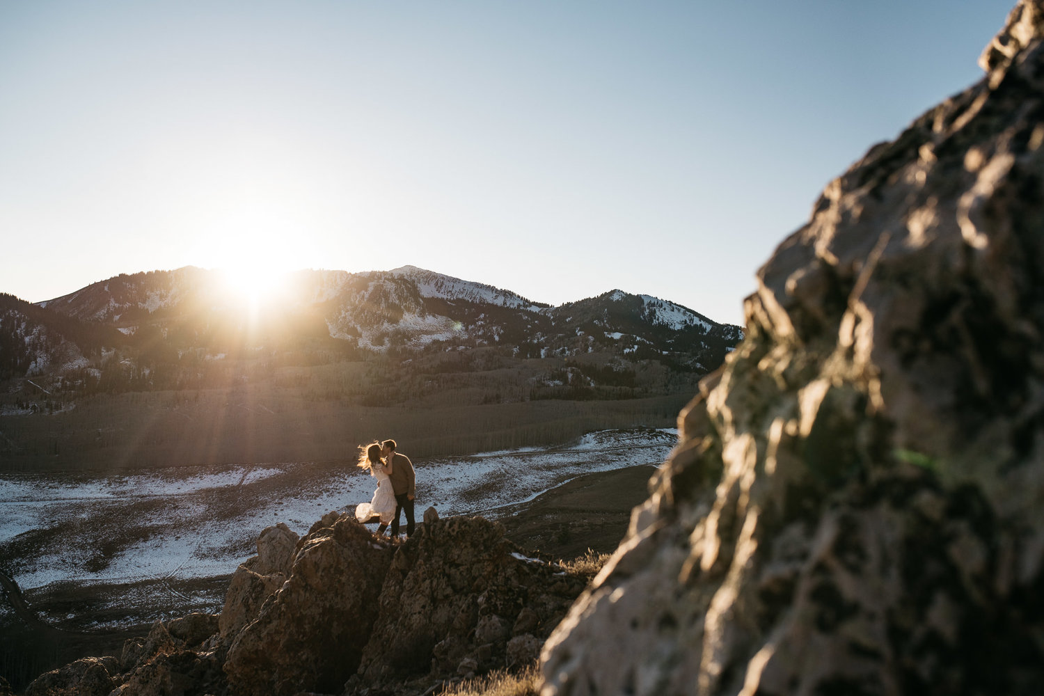 A couple kissing while standing on top of a cliff as the sun shines through from behind the mountains in the background