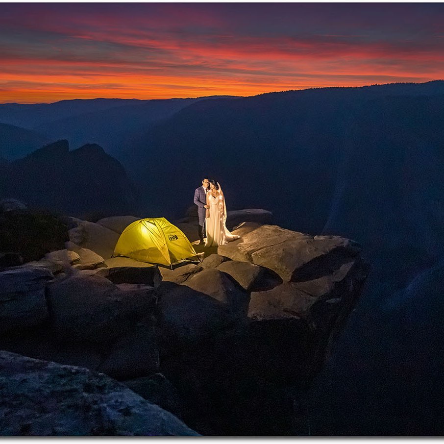 A bride and groom posing on top of a cliff alongside a camping tent with a sunset sky in the background