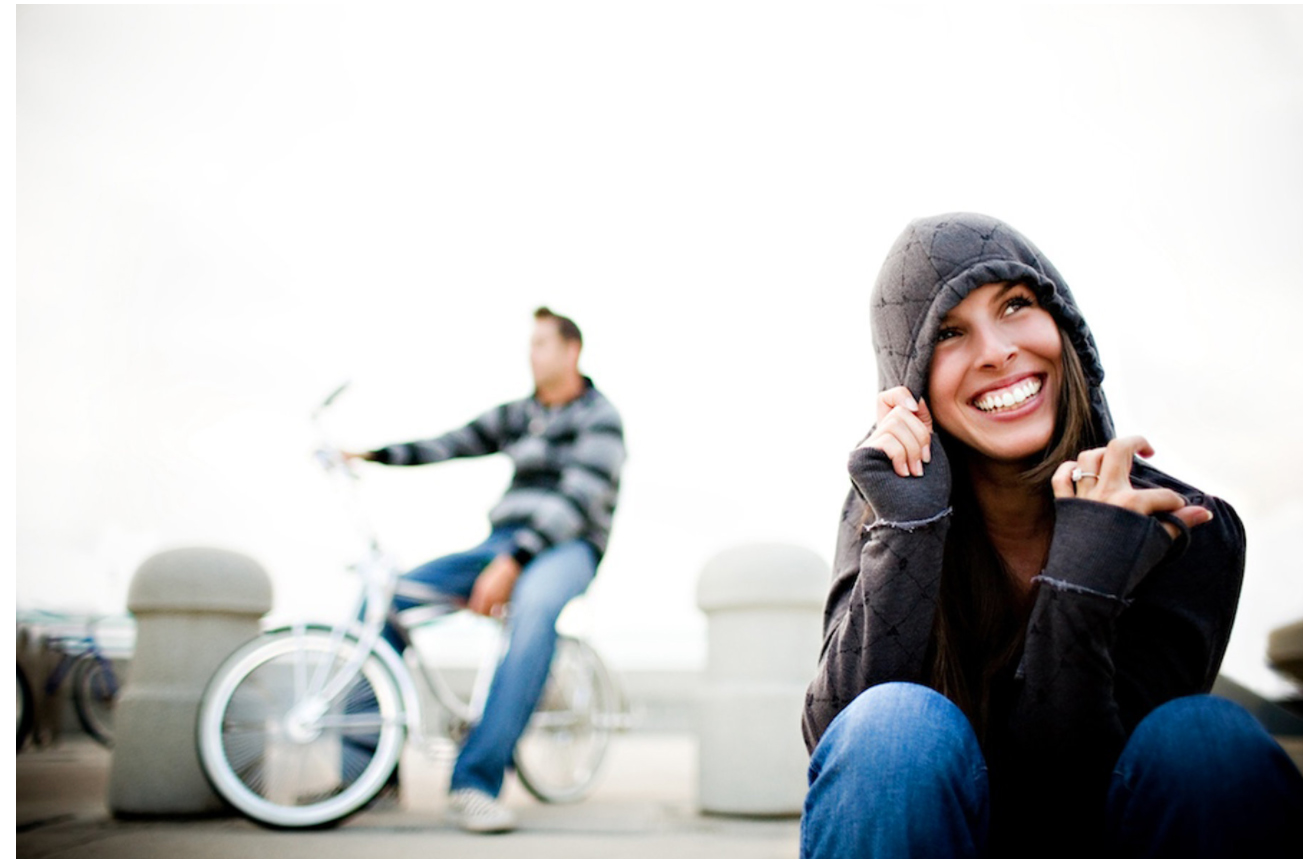 A girl posing wearing a hoodie with a guy sitting on a bicycle in the background