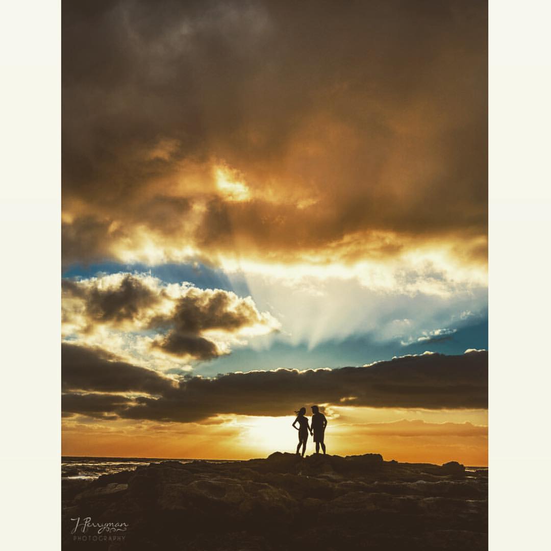A silhouette of a couple posing on a rocky beach with a sunset and stormy sky background 