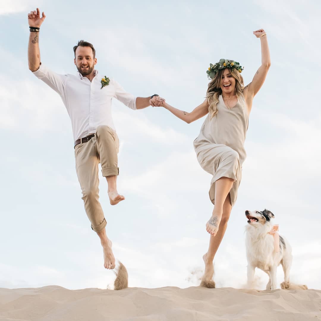 A bride and groom posing for a jump shot in a desert with their dog at their side (showcasing love is in the air)