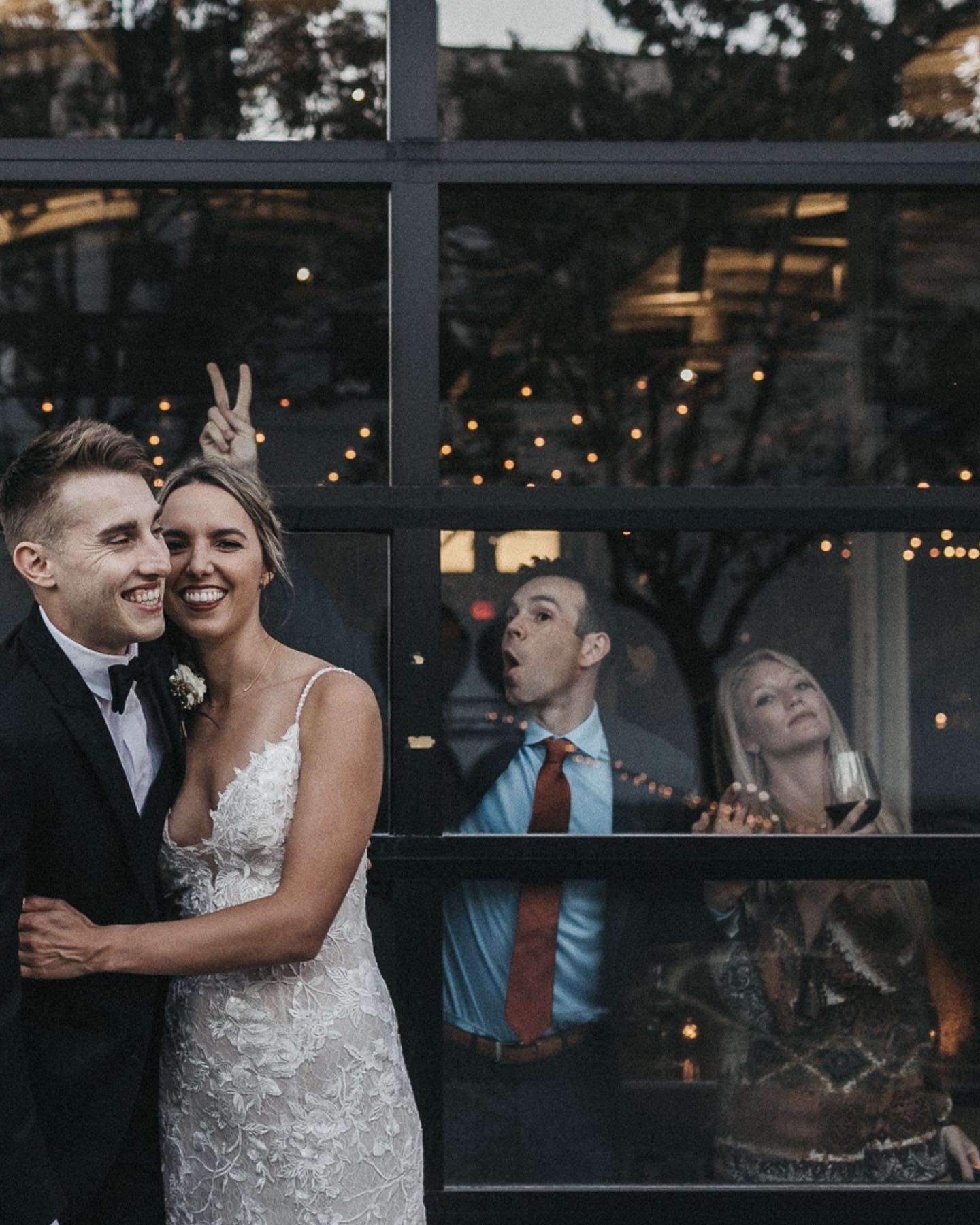a married couple is posing while their friends making funny gestures from the other side of a glass pane